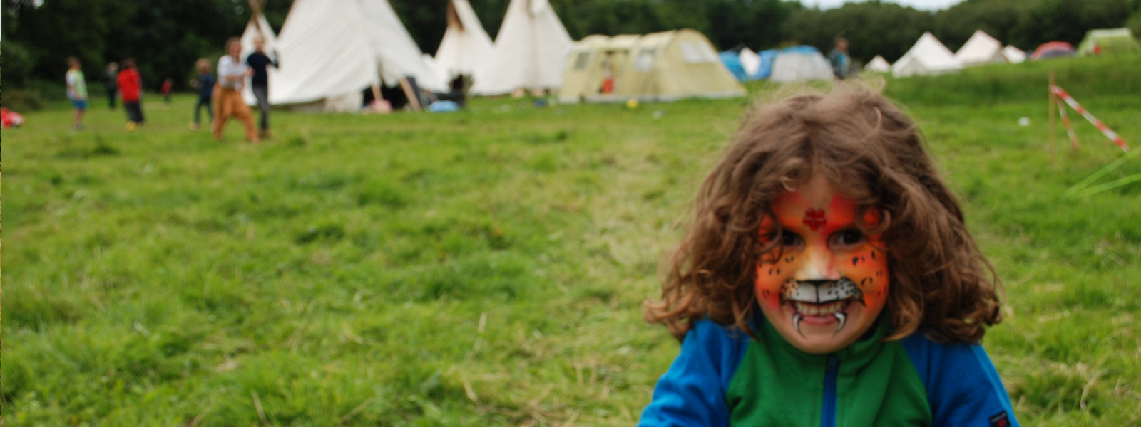 Small child with tiger make up standing in a green field in front of teepees